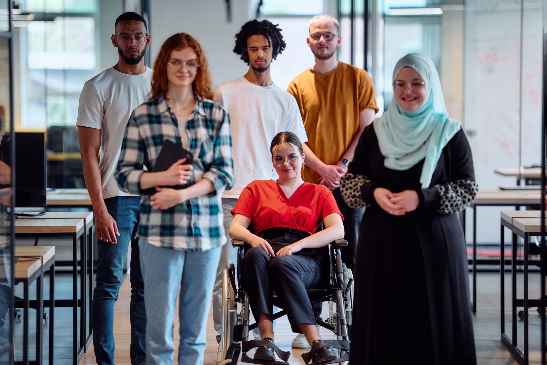 A diverse group of young business people walking a corridor in the glass-enclosed office of a modern startup, including a person in a wheelchair and a woman wearing a hijab